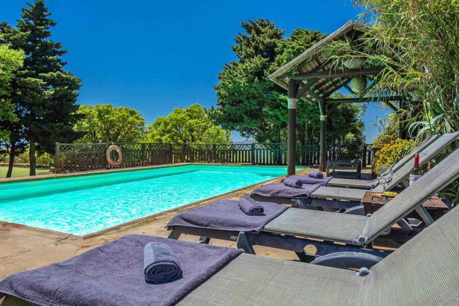 Piscina del Chalet La Ahumada con tumbonas, toallas, pérgola de madera y vegetación alrededor, bajo un cielo azul intenso.