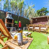 Primer plano de una botella de vino tinto y dos copas sobre una mesa de madera entre tumbonas en el jardín de Villa Verde, con barbacoa y mesa exterior al fondo.