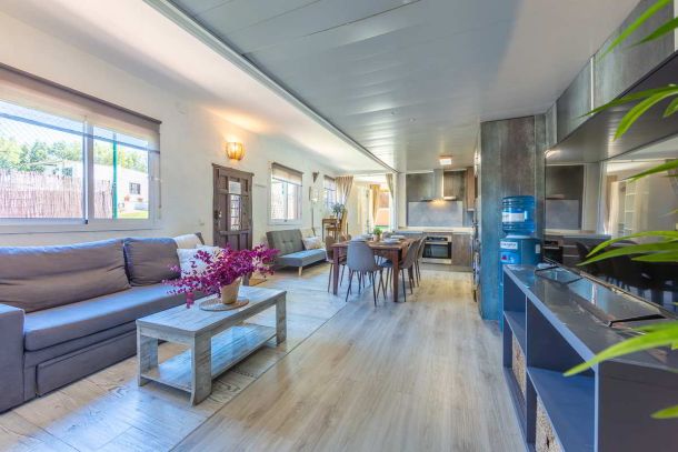 Salón-comedor con sofá gris, mesa de centro con flores fucsias, zona de comedor con mesa de madera y seis sillas, cocina integrada con muebles oscuros y electrodomésticos modernos, televisión sobre mueble azul y grandes ventanales que iluminan todo el ambiente.