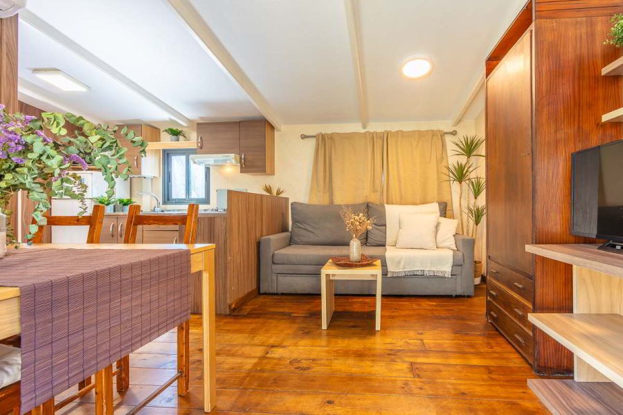 Salón de la casa Nepal en Villas La Ahumada, con suelo de madera, sofá gris con cojines blancos, mesa baja con jarrón de flores secas, comedor de madera con centro floral y cocina integrada al fondo en tonos cálidos