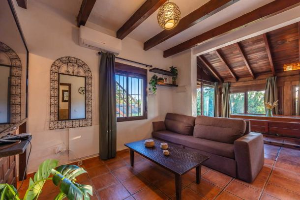Salón rústico de Villa Paraíso, Villas La Ahumada, Tarifa, con sofá marrón, mesa de madera oscura, espejo decorativo con marco de forja, techo de vigas y ventanales con vistas al jardín.