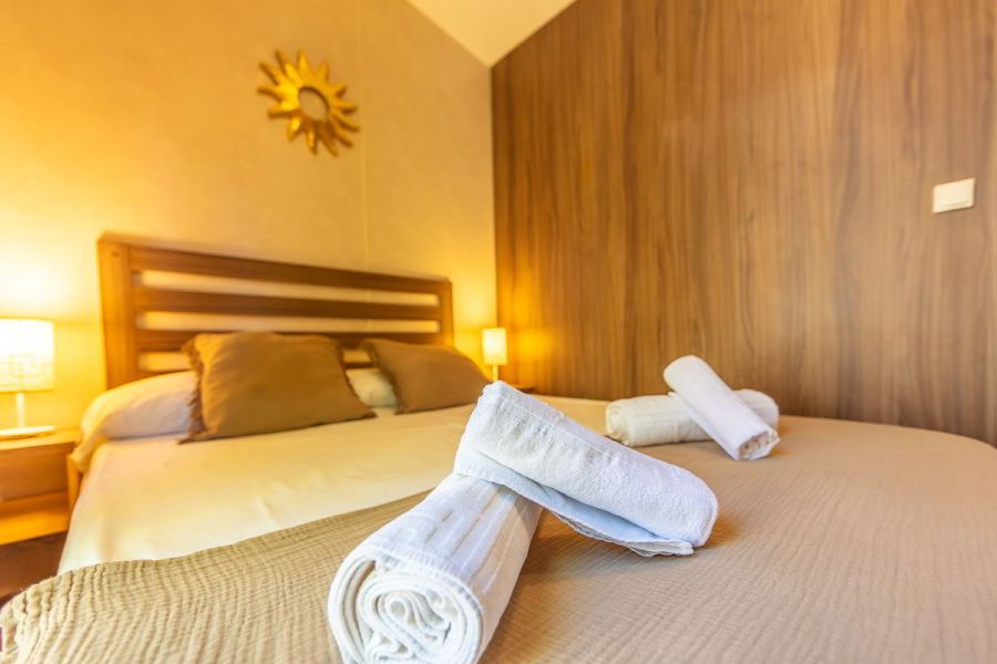 Detalle de la cama doble del dormitorio principal en la casa Nepal de Villas La Ahumada, con toallas enrolladas sobre la colcha beige, cojines marrones, cabecero de madera, lámparas encendidas y decoración dorada en forma de sol sobre la pared