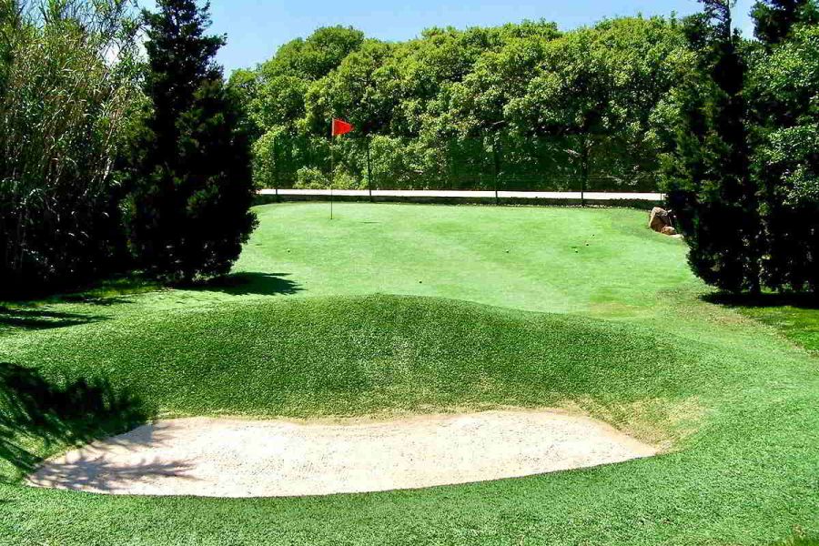 Campo de minigolf en Villa Paraíso, dentro del chalet La Ahumada, con bandera roja, césped cuidado y zona de arena rodeada de árboles.