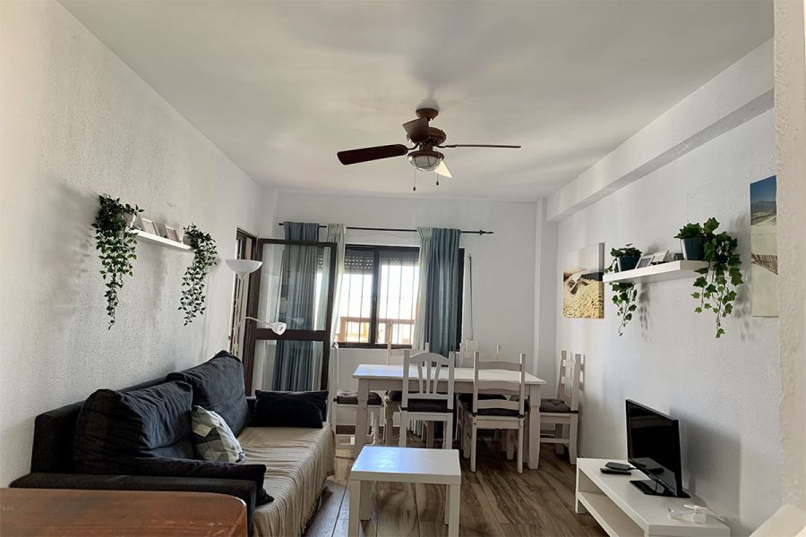 Sala de estar con sofá oscuro, lámpara blanca, TV y una cocina abierta moderna al fondo.