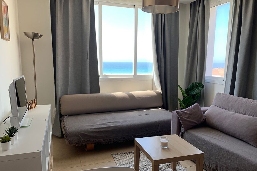 Salón con sofá gris, ventana grande y vistas al mar.