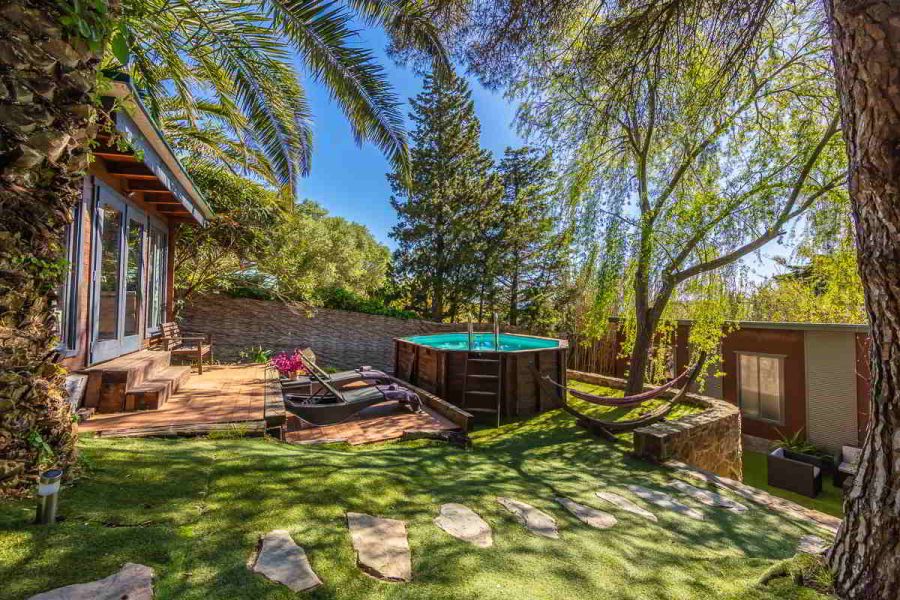 Imagen del jardín con piscina elevada de madera, tumbonas sobre tarima, una hamaca entre árboles y una cabaña de madera bajo las palmeras en el alojamiento en Tarifa.