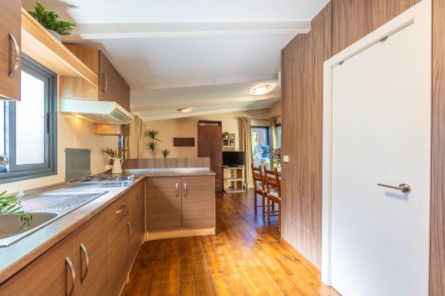 Vista desde el fondo de la cocina de la casa Nepal en Villas La Ahumada, con encimera en forma de L, muebles en madera, comedor al fondo con mesa y sillas, televisión, plantas decorativas y grandes ventanales con salida al jardín
