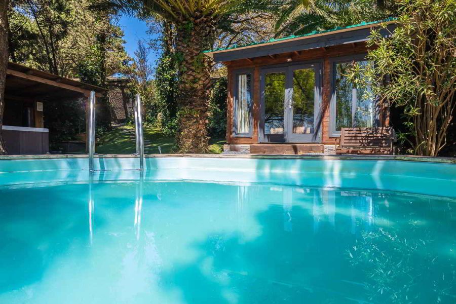 Vista de la piscina de la casa Nepal en Villas La Ahumada, rodeada de palmeras y vegetación frondosa, con escalerilla metálica, casita de madera con grandes ventanales al fondo y banco de madera junto a la entrada