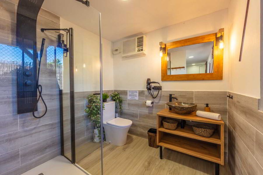 Cuarto de baño con ducha de cristal y columna negra, inodoro moderno, lavabo de piedra sobre mueble de madera con cestas, espejo con marco de madera, luz cálida, suelos efecto madera y detalles vegetales decorativos.
