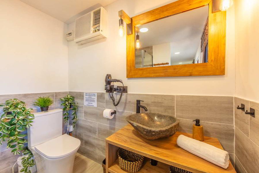 Detalle del baño con lavabo de piedra sobre encimera de madera, espejo con marco de madera e iluminación cálida, WC con plantas decorativas, secador de pelo y pared con azulejos grises estilo madera.